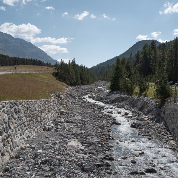 Fluss Ova dal Fuorn mit Steinmauern, Straße und Bergen im Hintergrund.