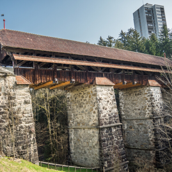 Gedeckte Holzbrücke in Rothenburg (Rotbach) mit Steinsäulen und Hochhaus im Hintergrund.