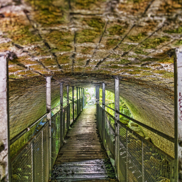 Überdachter Gang mit Steinwänden und Metallgeländer, der ins Helle führt.