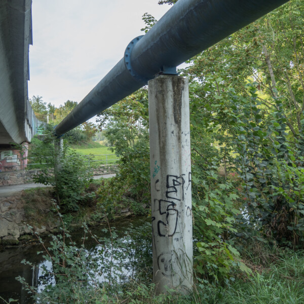 Betonpfeiler unter einer Brücke mit einer großen Rohrleitung, Graffiti und Vegetation.