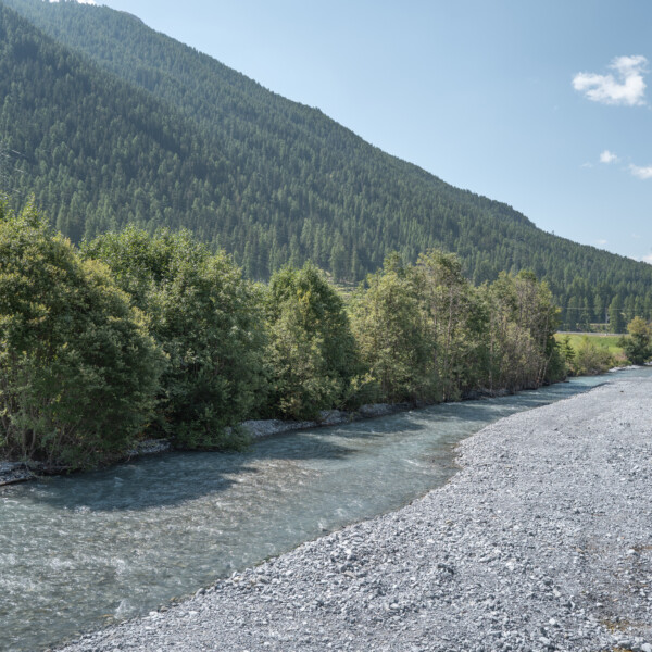 Fluss Spöl mit Kiesbett und bewaldetem Berghang unter blauem Himmel.