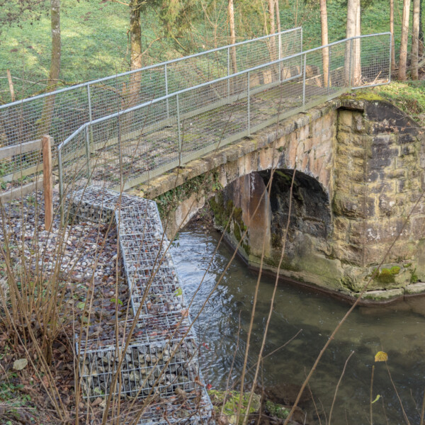 Steinbrücke mit Metallgeländer über einen kleinen Fluss. Uferbefestigung mit Steinkörben.