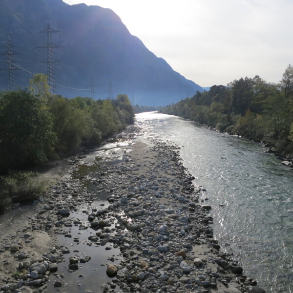 Flusslandschaft im Tessin mit Bergen und Strommasten.