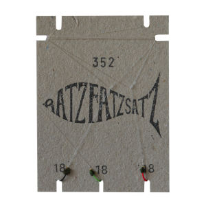 Auf strukturiertem Karton ist „RatzFatzSatz 352“ in Form eines Fisches abgebildet, an dessen Unterseite drei Knoten aus grünen, roten und schwarzen Fäden mit der Aufschrift „18“ angebracht sind.