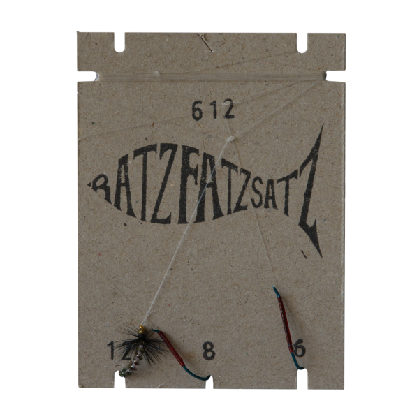 Ein fischförmiger Papphalter mit der Aufschrift „RatzFatzSatz 612 (Weiss)“ trägt darunter die Nummern 12, 8 und 6 und an drei Schnüren sind Weiss-Angelfliegen befestigt.