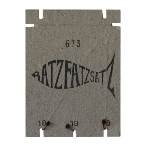 Ein fischförmiges Motiv auf braunem Karton trägt im Inneren den Schriftzug „RatzFatzSatz 673 (Grün, Rot & Violett)“, akzentuiert in Grün, Rot und Violett. Oben und unten sind die Zahlen 673 und 18 platziert; Fäden und Knoten heben drei untere Punkte hervor.