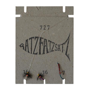 Eine braune Kartonpackung mit der Aufschrift „RatzFatzSatz 727 (Weiss, Pink & Violett)“ enthält zwei Angelfliegen (gekennzeichnet mit 16 und 7) mit einem fischförmigen Logo als Hinweis auf RatzFatzSatz-Authentizität. Eine Fliege ist in Pink und Violett gehalten.