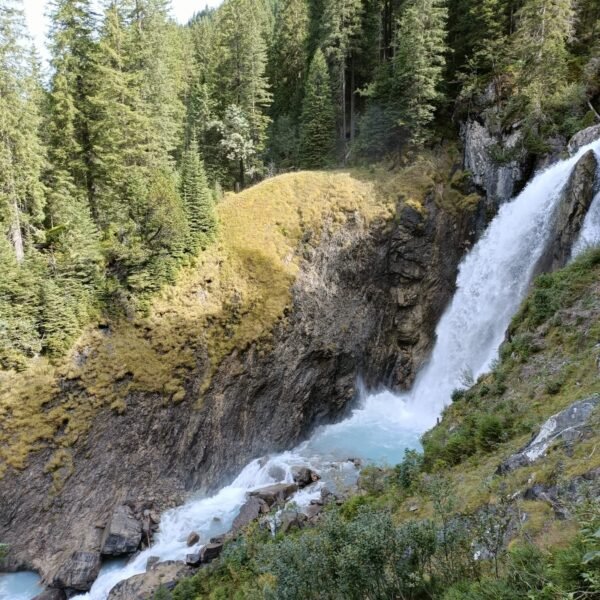 Ein Wasserfall stürzt eine felsige Klippe hinunter, die von dichten grünen Kiefern und grasbewachsenen Hängen umgeben ist, und am Fuße sammelt sich schäumendes blaues Wasser. Das Sonnenlicht erhellt Teile der Landschaft.