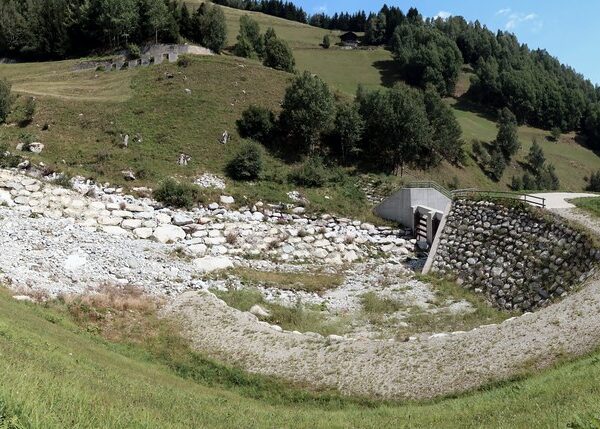 Acletta: Bachbett mit Steinen und einer Betonkonstruktion in einer grünen Landschaft.