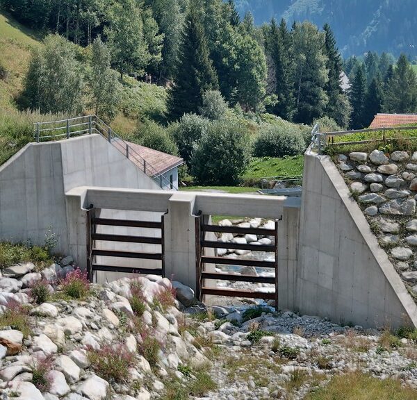 Acletta: Betonbauwerk mit Steinen und Gitter im Flussbett, umgeben von grüner Landschaft.