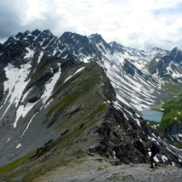 Wanderer auf dem Grat mit Blick auf den Älplisee und schneebedeckte Berge.