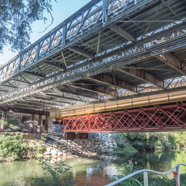 Alter Rhein Brücke: Stahlkonstruktion über Fluss mit Uferbewuchs und blauem Himmel.