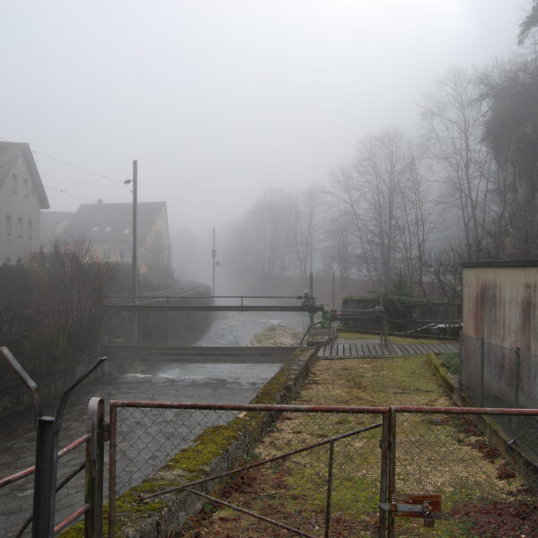Nebliger Blick auf den Augstbach mit Brücke und Uferbefestigung. Düstere Flusslandschaft.