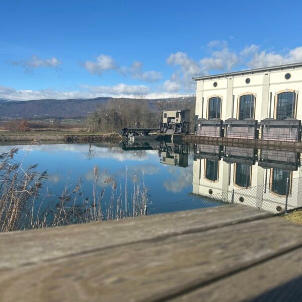 Ein cremefarbenes Gebäude mit gewölbten Fenstern spiegelt sich in einem ruhigen Gewässer unter strahlend blauem Himmel, mit Bergen und Schilf im Hintergrund und einer Holzfläche im Vordergrund.
