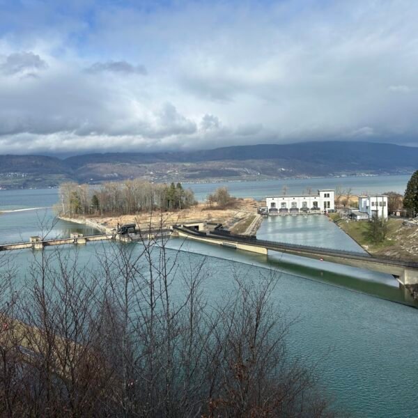 Eine malerische Ansicht eines Staudamms mit türkisfarbenem Wasser, Gebäuden und Bäumen vor einem großen See und entfernten Bergen unter einem bewölkten Himmel.