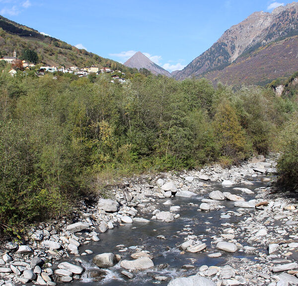 Fluss Brenno mit Felsen und Bäumen, im Hintergrund ein Dorf in den Bergen.