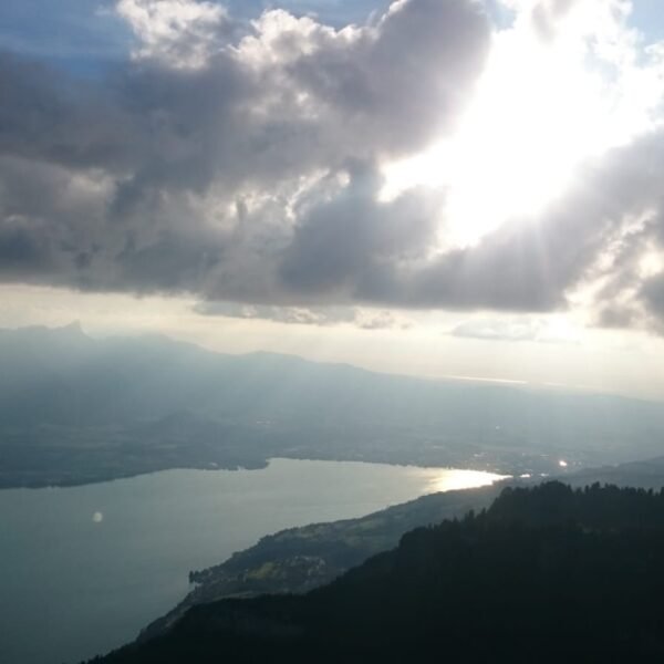 Ein malerischer Blick auf einen von Bergen umgebenen See unter einem dramatischen Himmel, wobei das Sonnenlicht durch dichte Wolken dringt und sich auf dem Wasser spiegelt.