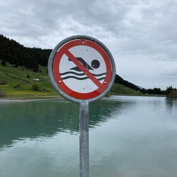 Am ruhigen Brigelsersee (Lag da Breil) steht ein rundes Schild mit einer roten diagonalen Linie, die ein Badesymbol durchkreuzt. Es weist darauf hin, dass das Baden verboten ist. Im Hintergrund sind grüne Hügel und ein bewölkter Himmel zu sehen.