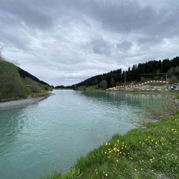 Ein ruhiger Fluss fließt unter einem bewölkten Himmel, gesäumt von grasbewachsenen Ufern mit gelben Blumen und Bäumen. Rechts befinden sich Tribünen am Wasser des Lag da Breil, im Hintergrund erhebt sich ein bewaldeter Hügel.