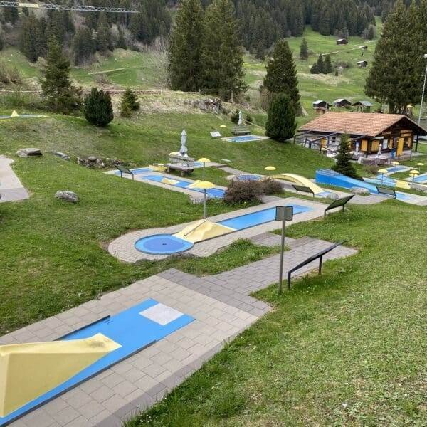 Minigolfplatz im Freien mit blauen und gelben Hindernissen in der Nähe des Lag da Breil, umgeben von grünem Gras, Bäumen und einem Holzgebäude im Hintergrund. Bänke und ein Spielplatz sind ebenfalls zu sehen.