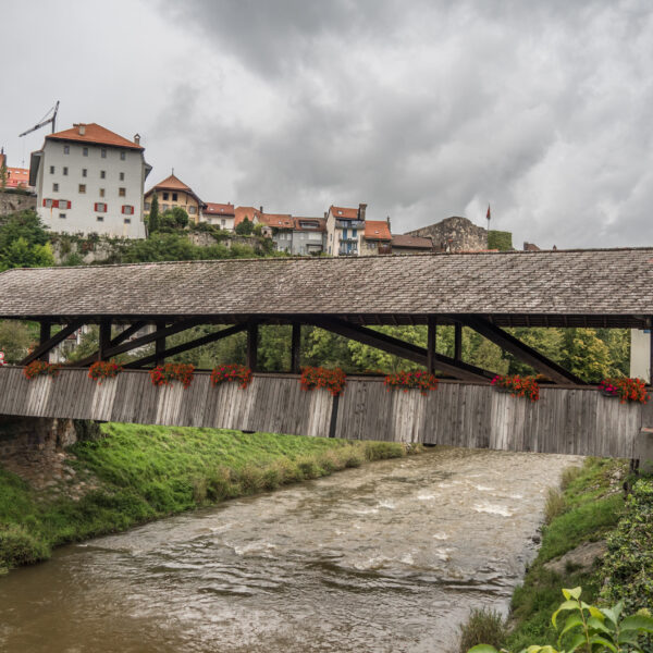 Gedeckte Holzbrücke über die Broye mit Blick auf mittelalterliche Gebäude und bewölkten Himmel.