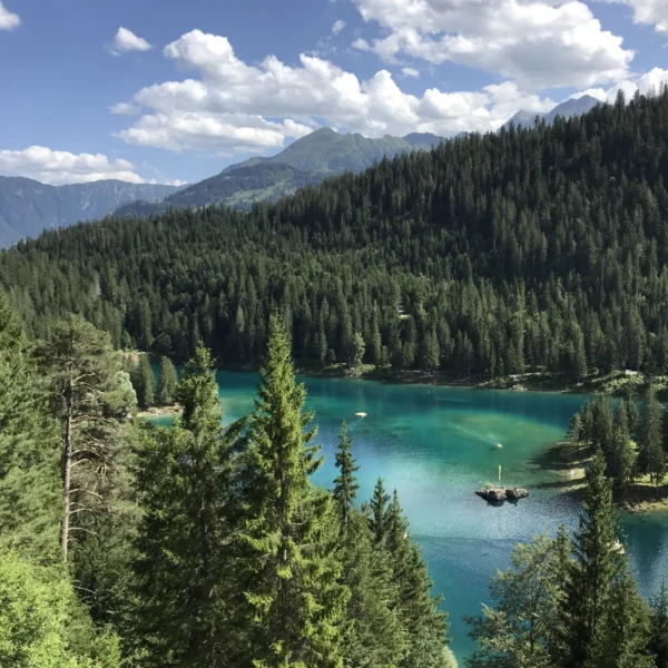 Ein türkisfarbener See, umgeben von dichten grünen Kiefernwäldern und Bergen unter einem teilweise bewölkten Himmel, wobei das Sonnenlicht Teile der Landschaft hervorhebt.