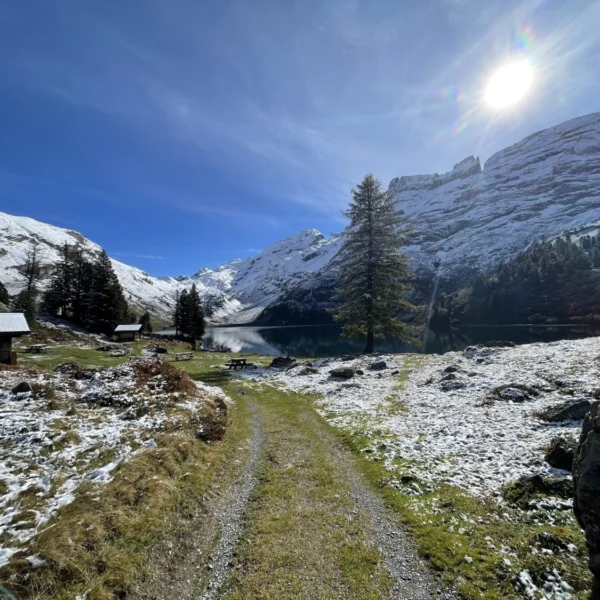 Ein Schotterweg führt an verschneitem Gras und kleinen Hütten vorbei zu einem ruhigen See, der von schneebedeckten Bergen und Bäumen umgeben ist und unter einem strahlend sonnigen Himmel steht.