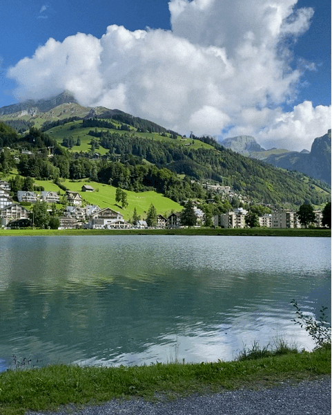 Eugenisee See mit Bergen und Häusern im Hintergrund. Ruhige Wasseroberfläche.