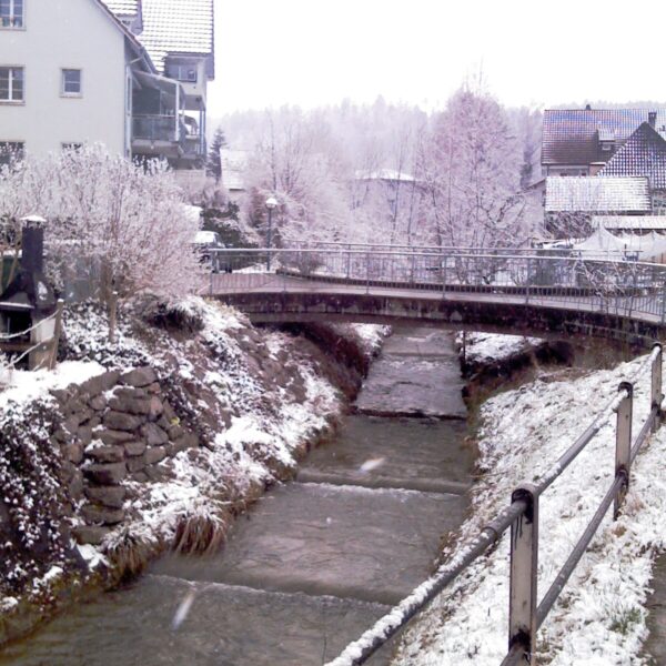 Winterliche Eulach: Bach unter Brücke mit Schnee, Häuser im Hintergrund.