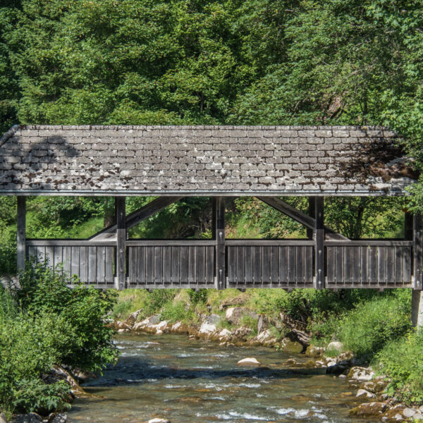 Überdachte Holzbrücke über einen Bach, umgeben von üppigem Grün.