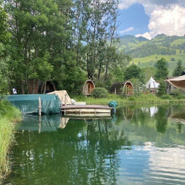 Eine ruhige Seelandschaft mit klarem Wasser, grünem Gras und Bäumen. Kleine Holzhütten und Zelte schmiegen sich in das Grün, mit Hügeln und Bergen im Hintergrund unter einem teilweise bewölkten Himmel.