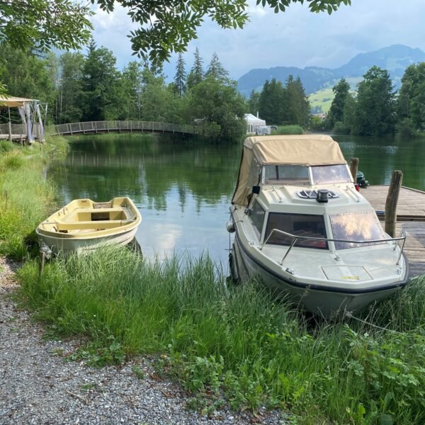Zwei kleine Boote liegen an einem Holzsteg an einem klaren See, der von üppigem Grün und Bäumen umgeben ist. Ein Kiesweg führt am Ufer entlang, und im Hintergrund überquert ein Holzsteg den See. In der Ferne erheben sich Berge.