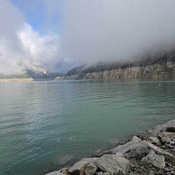 Ein ruhiger, grünlich-blauer See – der Göscheneralpsee – ist von felsigen Ufern und steilen Klippen gesäumt. Tief hängende Wolken und Nebel verdecken die Berge im Hintergrund teilweise. Ein paar kleine Eisberge schwimmen auf dem Wasser.