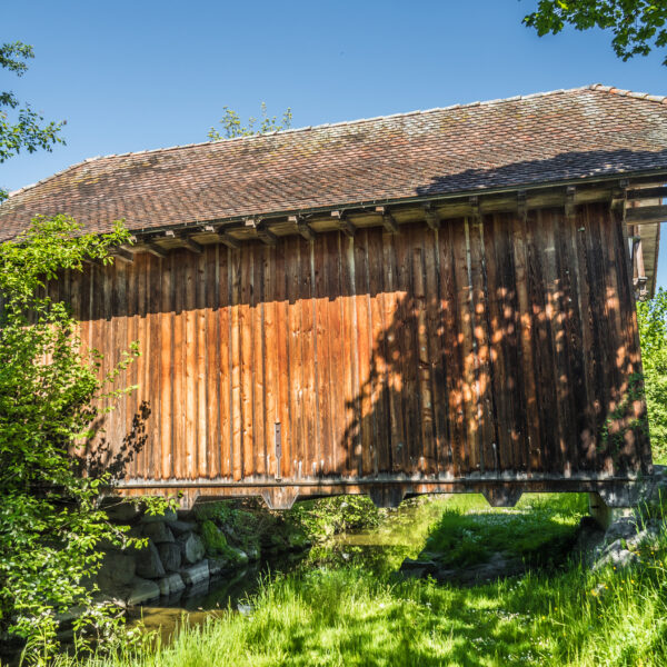 Grosse Aa: Überdachte Holzbrücke über einen Bachlauf, umgeben von Grün