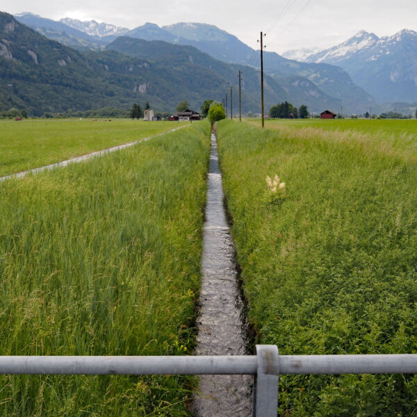 Hauptkanal-Wasserweg durch grüne Felder mit Bergen im Hintergrund.