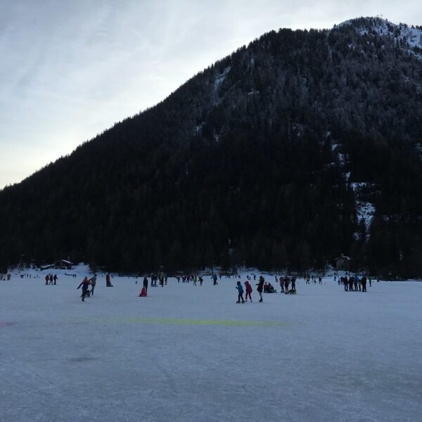 Lac de Champex im Winter: Menschen eislaufen auf dem gefrorenen See vor einem bewaldeten Berg.
