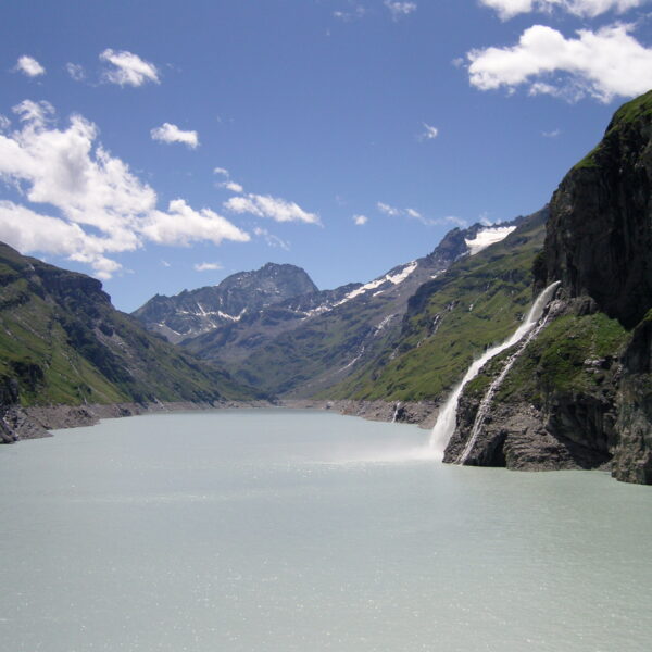 Lac de Mauvoisin mit Wasserfall und Bergen unter blauem Himmel.