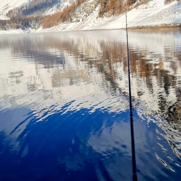 Eine Angelrute erstreckt sich über das ruhige, klare Wasser des Lac de Tseuzier, in dem sich verschneite Berge und Bäume unter einem strahlend blauen Himmel spiegeln. Sanfte Wellen kreuzen die Oberfläche, während herbstliche Bäume der Schneelandschaft leuchtende Farben verleihen.