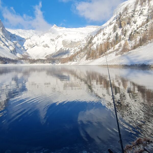 Eine ruhige, schneebedeckte Berglandschaft am Lac de Tseuzier in der Schweiz spiegelt sich in einem ruhigen Bergsee unter blauem Himmel, mit einer Angelrute am felsigen Ufer im Vordergrund.