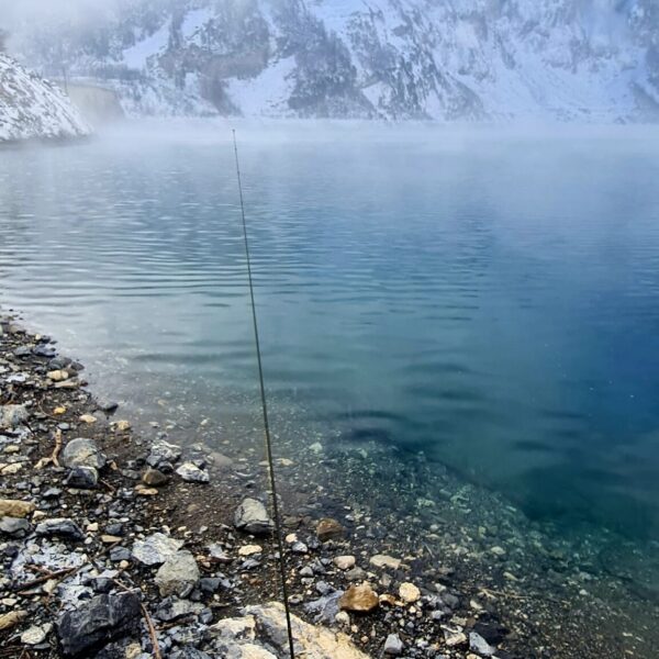 Eine Angelrute steht aufrecht an einem felsigen Ufer am klaren, ruhigen Wasser des Lac de Tseuzier. Im Hintergrund sind schneebedeckte Berge und Nebel zu sehen, die teilweise von Wolken umhüllt sind.