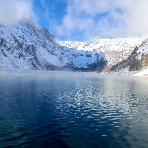 In einem klaren Bergsee spiegeln sich schneebedeckte Gipfel und Kiefern unter einem teilweise bewölkten blauen Himmel, und von der Wasseroberfläche steigt Nebel auf - eine ruhige Szene, die an den Lac de Tseuzier erinnert.