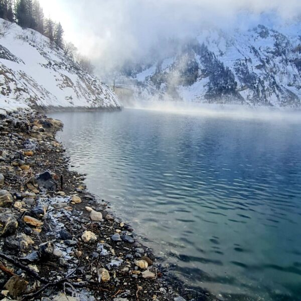 Felsiges Seeufer mit klarem Wasser im Vordergrund am Lac de Tseuzier, schneebedeckte, teilweise von Nebel und Wolken verhüllte Berge im Hintergrund unter blauem Himmel. Lichte Bäume säumen die linke Seite - ein einladender Ort für Wanderfreunde.