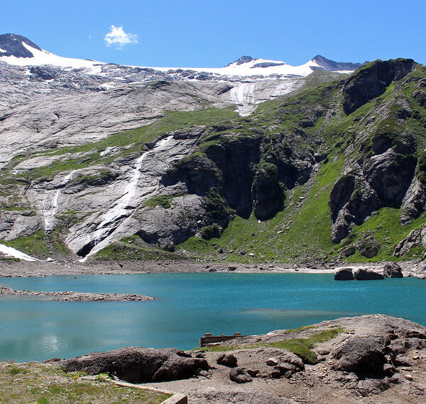 Türkisfarbener Lago del Zött vor schneebedeckten Bergen und grünen Hängen.