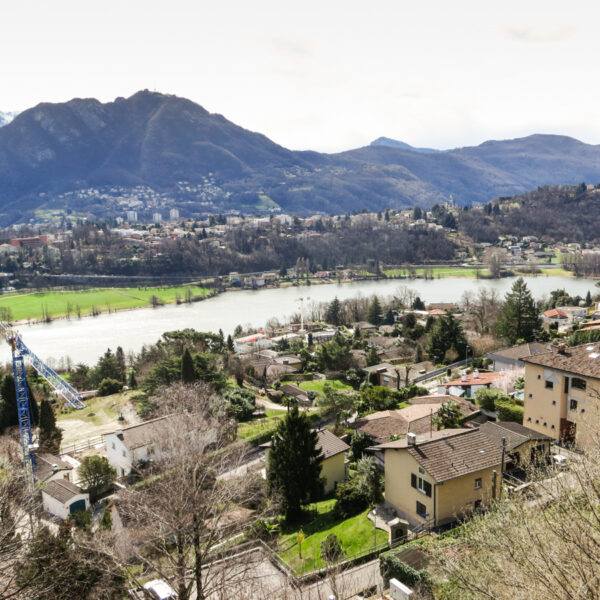 Blick auf den Lago di Muzzano mit Bergen und Häusern im Tessin, Schweiz.