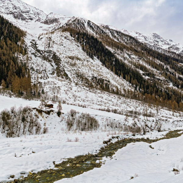 Winterlandschaft mit schneebedeckten Bergen und einem Bach. Lonza im Hintergrund.