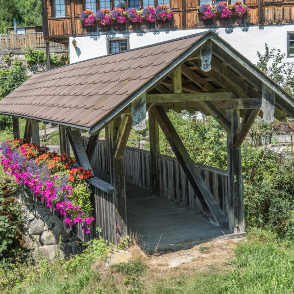 Überdachte Holzbrücke mit Blumenkästen in Lütisbach. Die Brücke führt über einen Bach.