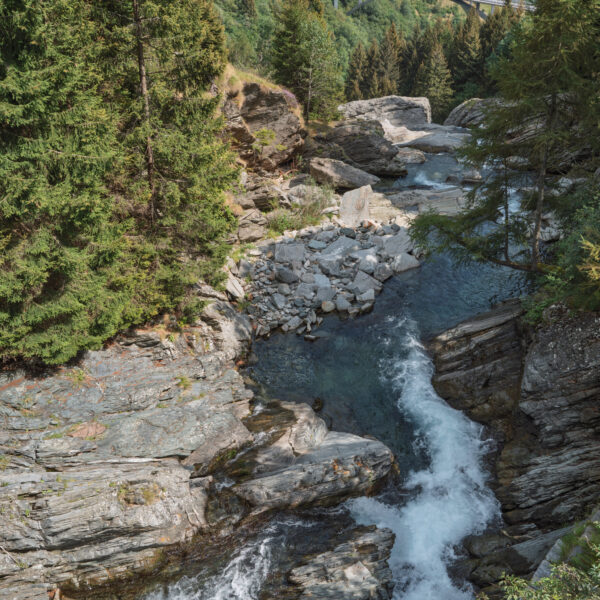 Moesa Fluss mit Kaskaden, Felsen und Bäumen. Eine Brücke ist im Hintergrund zu sehen.