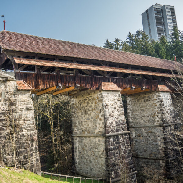 Gedeckte Holzbrücke in Rothenburg (Rotbach) mit Steinsäulen und Hochhaus im Hintergrund.