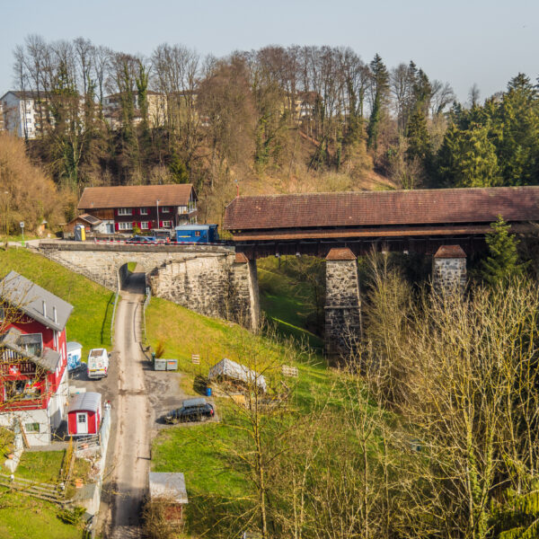 Rotbach (Rothenburg) mit gedeckter Holzbrücke und Häusern in ländlicher Umgebung.