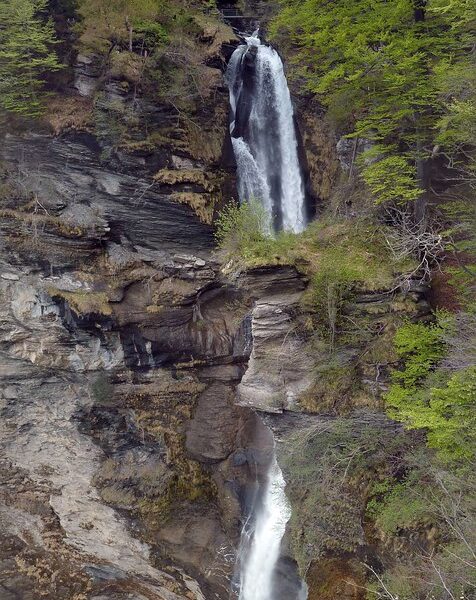 Rychenbachfall: Mehrstufiger Wasserfall in felsiger Landschaft mit Bäumen und einem Haus im Hintergrund.